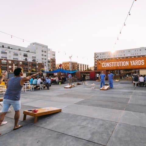 Rooftop courtyard with picnic tables and group playing cornhole under string lights with Constitution Yards sign