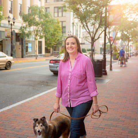 A woman smiling and walking her dog along a cobblestone sidewalk next to a city street