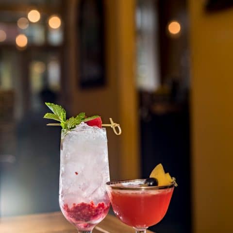 A small pink cocktail next to a tall white drink with crushed fruit in the bottom, both adorned with fruit garnish