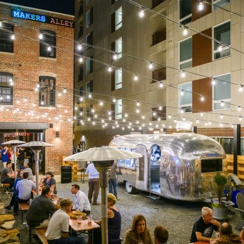 Makers Alley in the evening with outdoor picnic tables, an old metal camper, and a garage door entrance into a bar