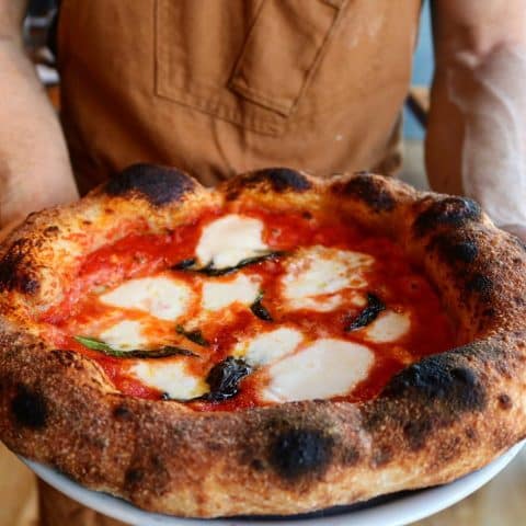 Person holding a Margherita pizza with mozzarella cheese and thick crust on a platter